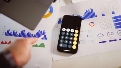 An overhead view of a desk where a hand holding a pencil rests on printed bar charts and pie charts, beside a smartphone showing an open calculator app.