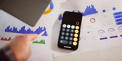 An overhead view of a desk where a hand holding a pencil rests on printed bar charts and pie charts, beside a smartphone showing an open calculator app.