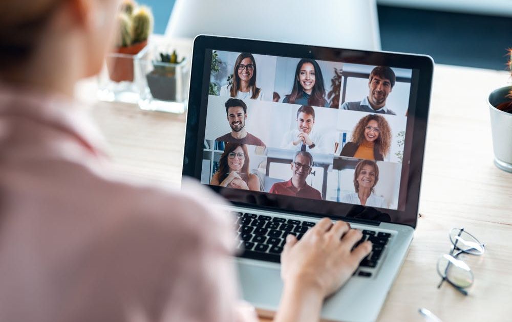 An over-the-shoulder view of a woman participating in a video conference call on her laptop, displaying a grid of nine smiling, diverse colleagues.