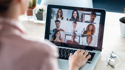 An over-the-shoulder view of a woman participating in a video conference call on her laptop, displaying a grid of nine smiling, diverse colleagues.