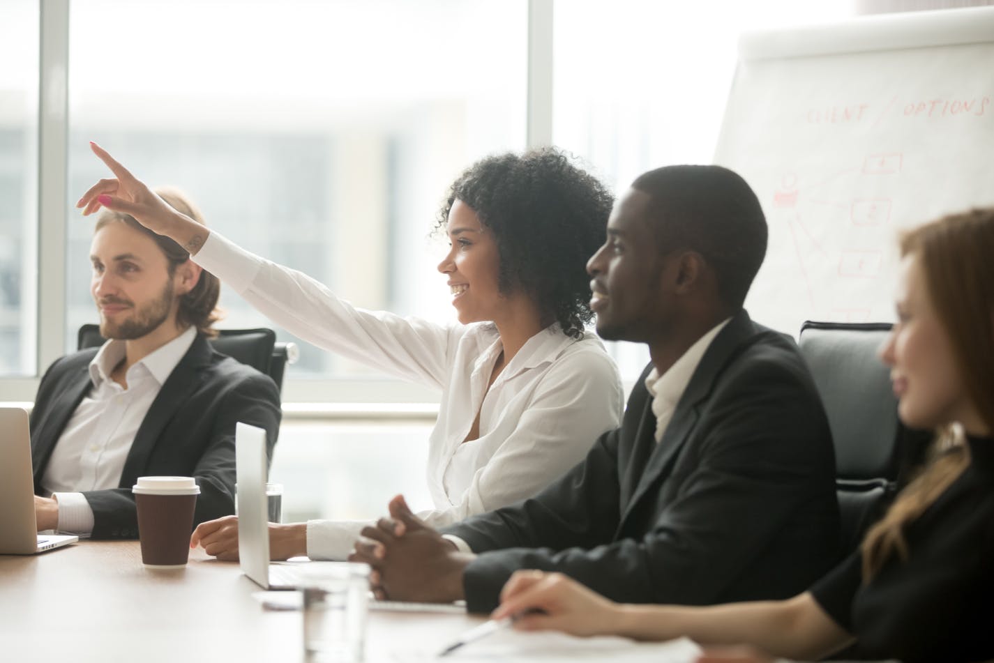 Een lachende Zwarte vrouw die haar hand opsteekt om een vraag te stellen of een bijdrage te leveren tijdens een zakelijke vergadering, zittend aan een vergadertafel naast diverse collega's.