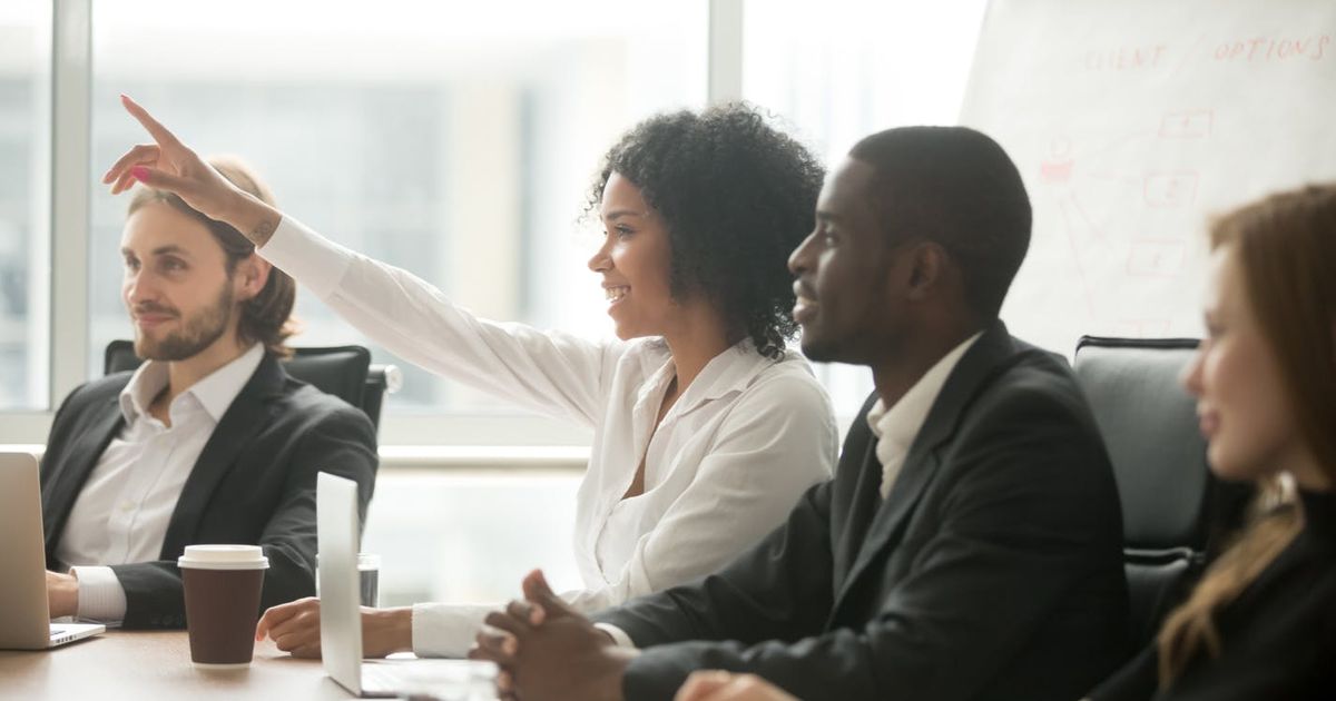 A smiling Black woman raising her hand to ask a question or contribute during a business meeting, sitting at a conference table alongside diverse colleagues.