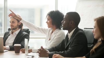 A smiling Black woman raising her hand to ask a question or contribute during a business meeting, sitting at a conference table alongside diverse colleagues.
