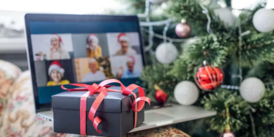 A wrapped gift box sits on a laptop in front of a decorated Christmas tree, while a video call with several people wearing Santa hats appears on the screen, suggesting a virtual holiday celebration.
