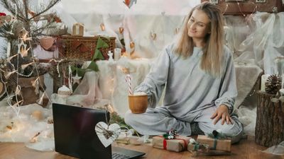 A smiling woman in grey pajamas sitting cross-legged on the floor, holding a mug with a candy cane while looking at an open laptop in a festively decorated holiday room.