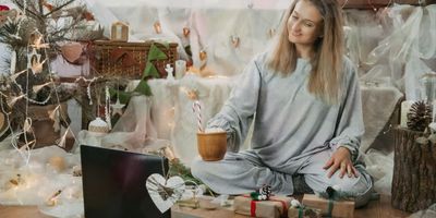 A smiling woman in grey pajamas sitting cross-legged on the floor, holding a mug with a candy cane while looking at an open laptop in a festively decorated holiday room.