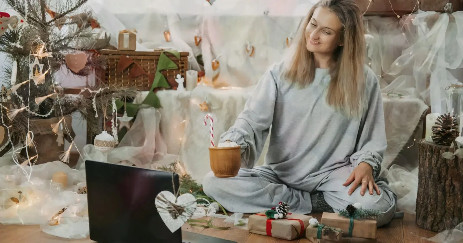 A smiling woman in grey pajamas sitting cross-legged on the floor, holding a mug with a candy cane while looking at an open laptop in a festively decorated holiday room.