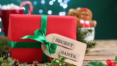 A close-up of a red Christmas gift wrapped with a green satin ribbon and a tag that reads "SECRET SANTA," set against a blurred holiday background.