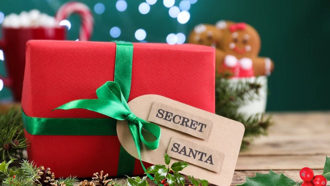 A close-up of a red Christmas gift wrapped with a green satin ribbon and a tag that reads "SECRET SANTA," set against a blurred holiday background.