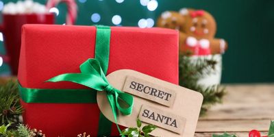 A close-up of a red Christmas gift wrapped with a green satin ribbon and a tag that reads "SECRET SANTA," set against a blurred holiday background.