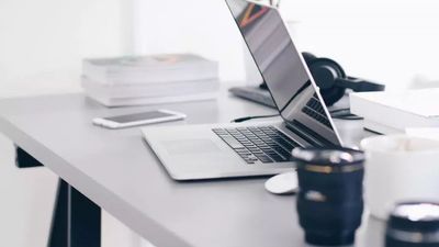A modern, minimalist workspace featuring an open laptop, a smartphone, a stack of books, and a pair of headphones on a clean grey desk.