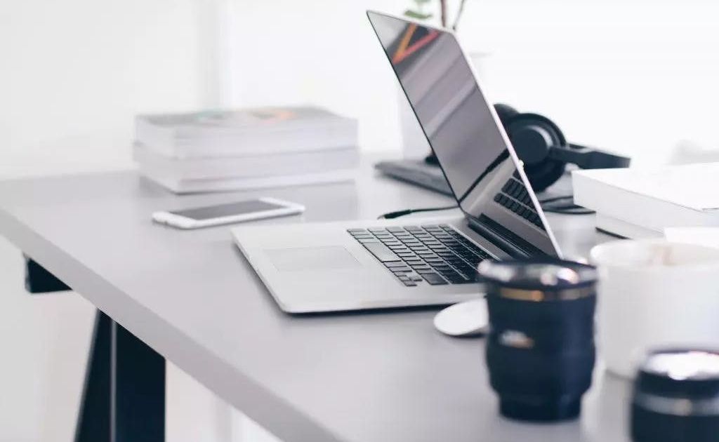 A modern, minimalist workspace featuring an open laptop, a smartphone, a stack of books, and a pair of headphones on a clean grey desk.