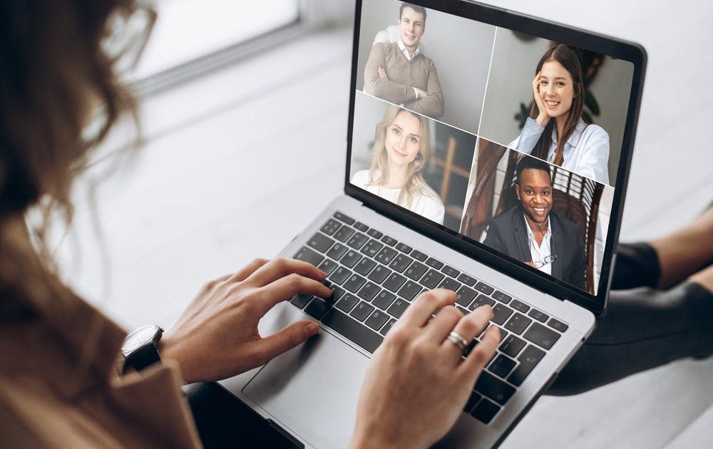 An over-the-shoulder view of a woman participating in a video conference call on her laptop with four smiling, diverse colleagues displayed on the screen.