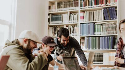 A group of young male students laughing together while gathering around a laptop to study in a library or classroom filled with books.