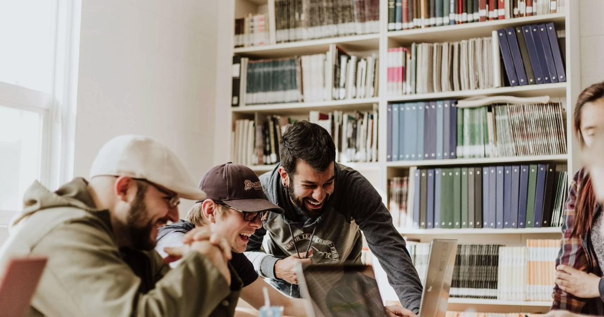A group of young male students laughing together while gathering around a laptop to study in a library or classroom filled with books.