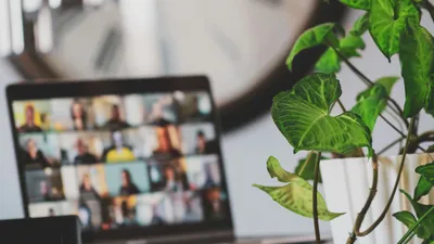A laptop on a desk showing a video call grid of participants, slightly out of focus behind a green houseplant in a white pot, with a large wall clock in the background.