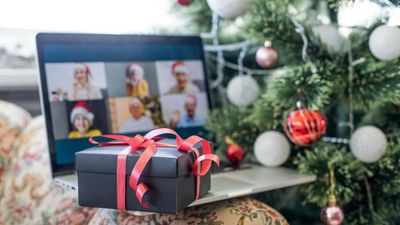 A black gift box wrapped with a red ribbon sitting on a laptop, which displays a virtual holiday group video call next to a decorated Christmas tree.