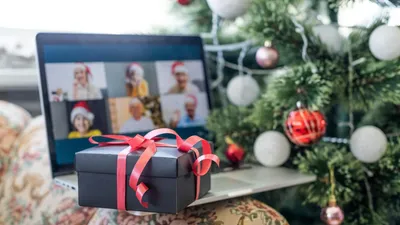 Une boîte cadeau noire ornée d'un ruban rouge posée sur un ordinateur portable, affichant un appel vidéo de groupe virtuel pour les fêtes, à côté d'un sapin de Noël décoré.