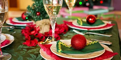 A holiday dinner table laid for a group, with white scalloped plates on red and dark green napkins, a red glitter ornament resting on the nearest setting, filled champagne glasses, and curled red ribbon running down a green damask tablecloth in front of blurred holiday lights.