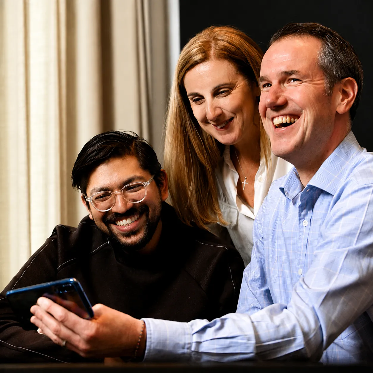 A group of three adults smiling and laughing together while looking at a smartphone, suggesting a fun shared activity or game.