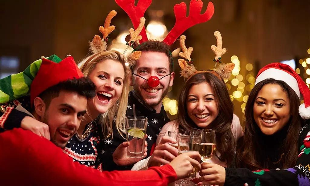 A joyful group of five friends smiling and clinking glasses together at a Christmas party, wearing festive attire like reindeer antlers, a Santa hat, an elf hat, and holiday sweaters.