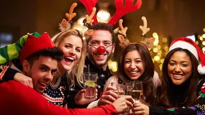 Un groupe joyeux de cinq amis souriant et trinquant ensemble lors d'une fête de Noël, portant des tenues festives comme des bois de renne, un bonnet de Père Noël, un chapeau de lutin et des pulls de Noël.