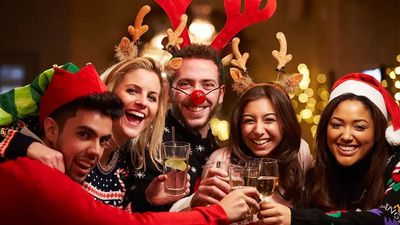 A joyful group of five friends smiling and clinking glasses together at a Christmas party, wearing festive attire like reindeer antlers, a Santa hat, an elf hat, and holiday sweaters.