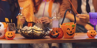 A close-up of a wooden table filled with Halloween decorations, popcorn in a spiderweb bowl, and small pumpkin buckets, with people in costumes sitting blurred in the background.