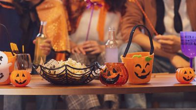 A close-up of a wooden table filled with Halloween decorations, popcorn in a spiderweb bowl, and small pumpkin buckets, with people in costumes sitting blurred in the background.