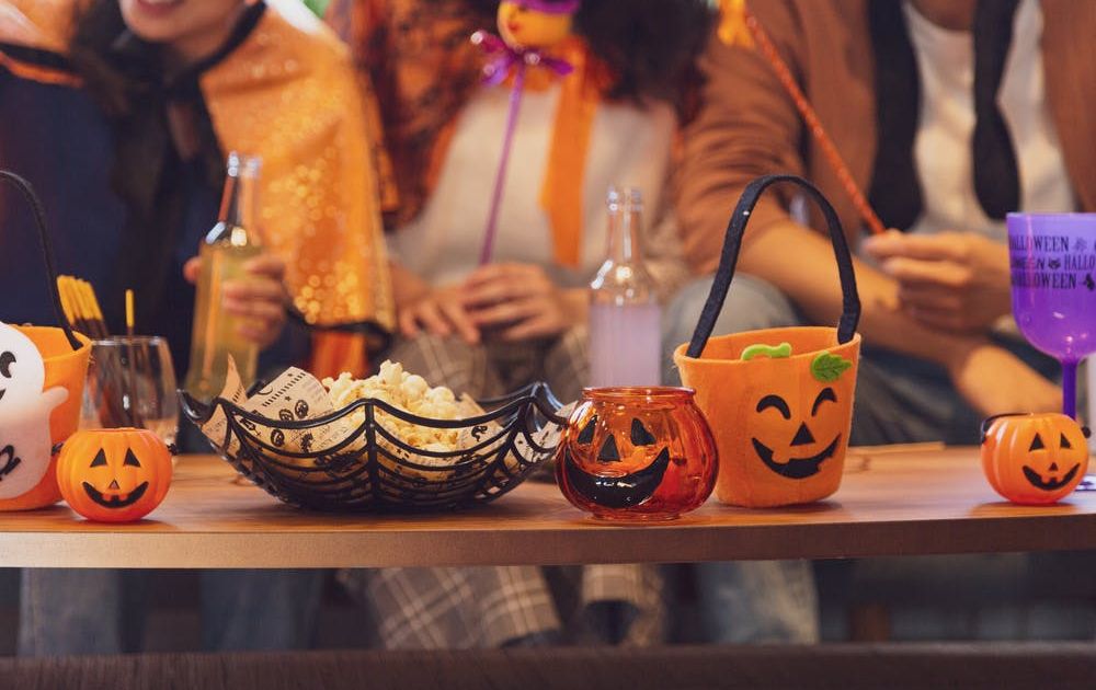 A close-up of a wooden table filled with Halloween decorations, popcorn in a spiderweb bowl, and small pumpkin buckets, with people in costumes sitting blurred in the background.