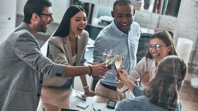 A high-angle view of a diverse group of five smiling colleagues clinking champagne flutes together to celebrate a success in their office.
