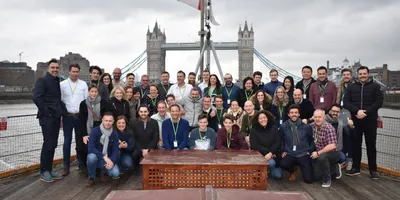 Een grote groep glimlachende mensen die poseren voor een foto op het dek van een boot op de Theems, met de beroemde Tower Bridge van Londen op de achtergrond onder een bewolkte hemel.