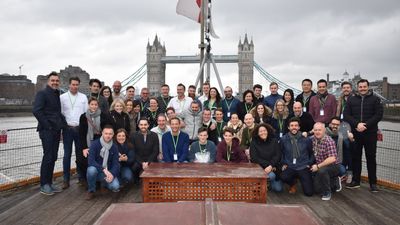 A large group of people smiling and posing for a photo on the deck of a boat on the River Thames, with the iconic Tower Bridge in London in the background under an overcast sky.
