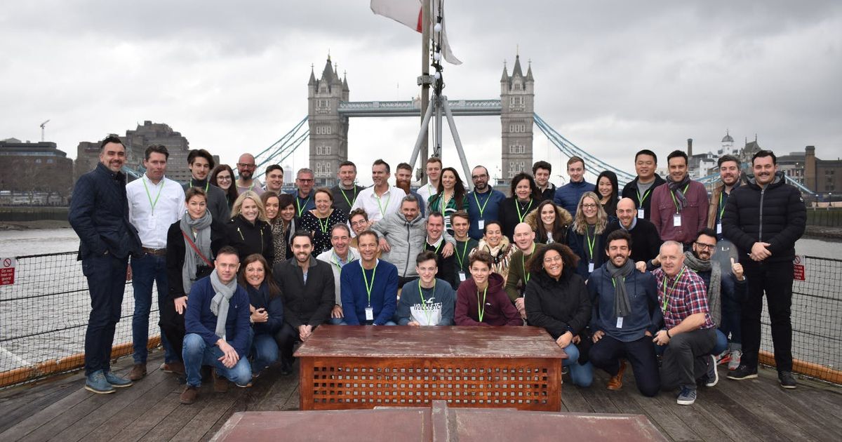 A large group of people smiling and posing for a photo on the deck of a boat on the River Thames, with the iconic Tower Bridge in London in the background under an overcast sky.
