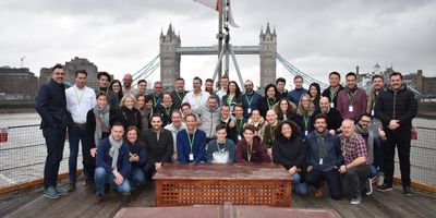 A large group of people smiling and posing for a photo on the deck of a boat on the River Thames, with the iconic Tower Bridge in London in the background under an overcast sky.