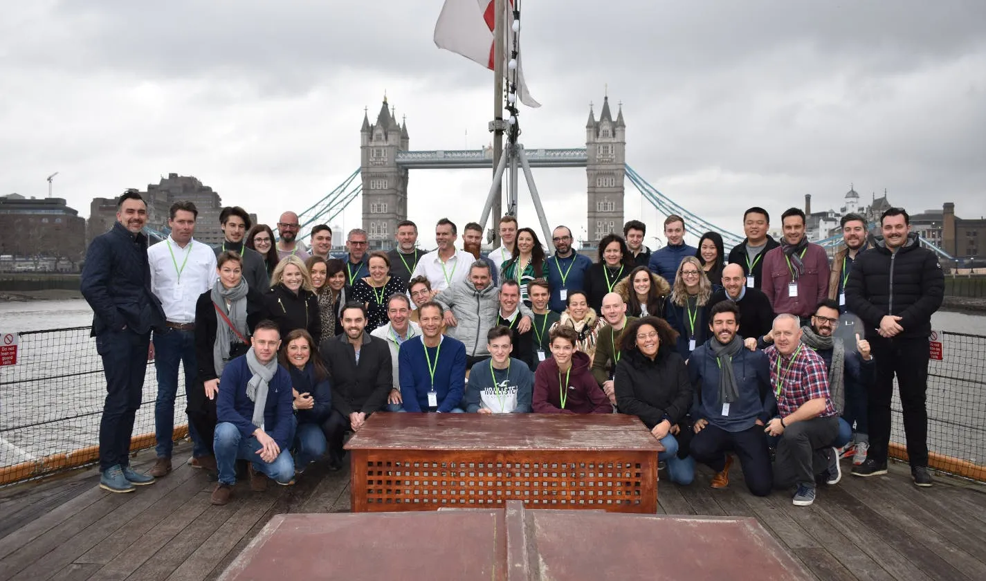 Een grote groep glimlachende mensen die poseren voor een foto op het dek van een boot op de Theems, met de beroemde Tower Bridge van Londen op de achtergrond onder een bewolkte hemel.