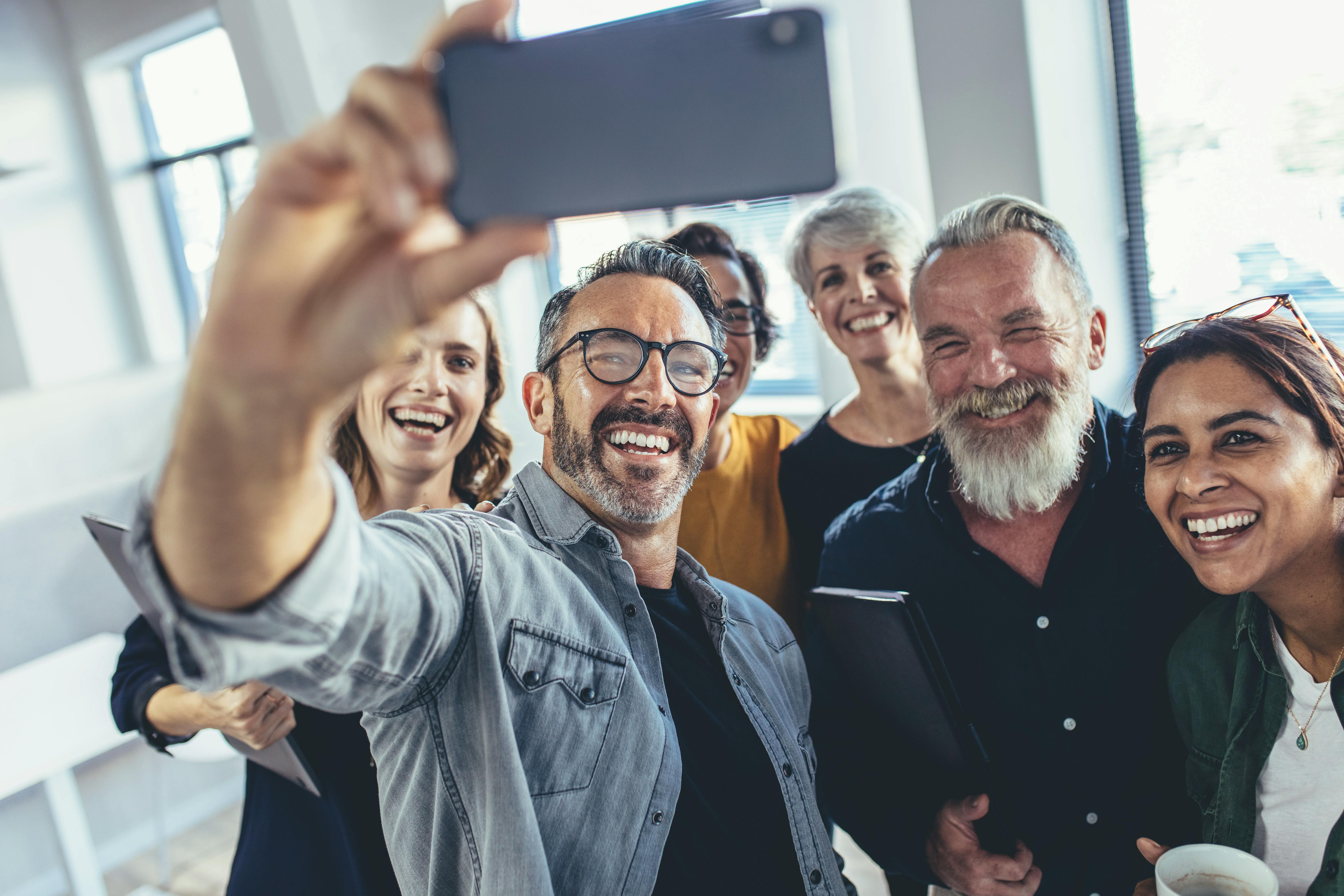 Een glimlachende, diverse groep volwassen collega's die samen in een licht kantoor bijeen zijn, lachend terwijl een man een smartphone omhooghoudt om een groepsselfie te maken.