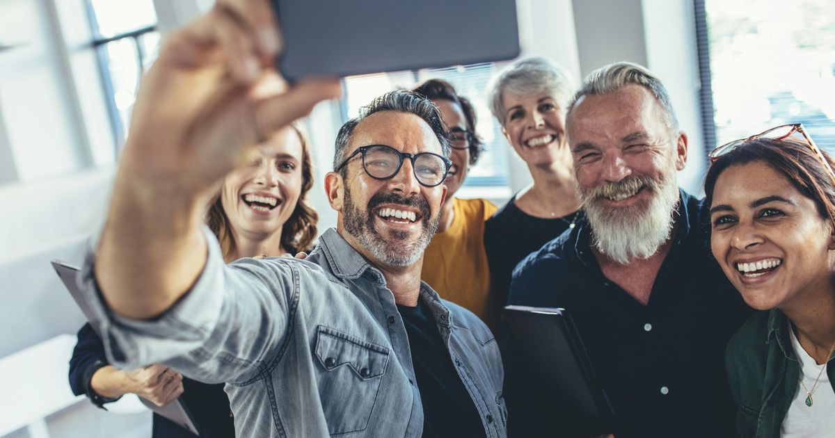 A smiling, diverse group of adult colleagues gathered together in a bright office, laughing as one man holds up a smartphone to take a group selfie.