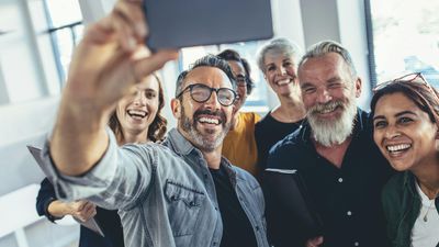A smiling, diverse group of adult colleagues gathered together in a bright office, laughing as one man holds up a smartphone to take a group selfie.