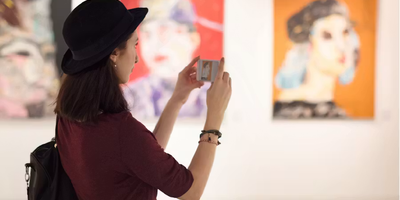 Vue de profil d'une femme portant un chapeau melon noir, utilisant son smartphone pour photographier un tableau coloré dans une galerie d'art.