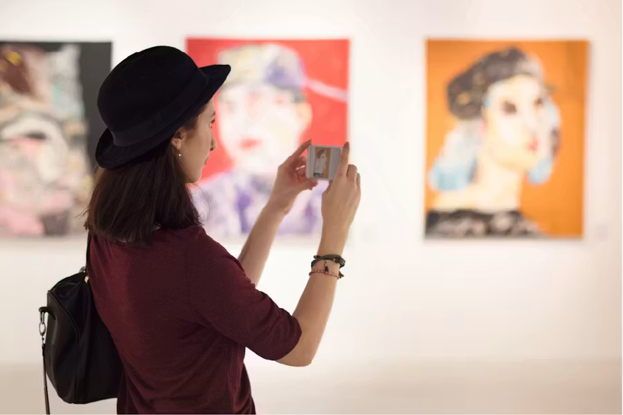 A side view of a woman wearing a black bowler hat, using her smartphone to take a photo of a colorful painting in an art gallery.