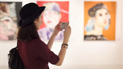 A side view of a woman wearing a black bowler hat, using her smartphone to take a photo of a colorful painting in an art gallery.