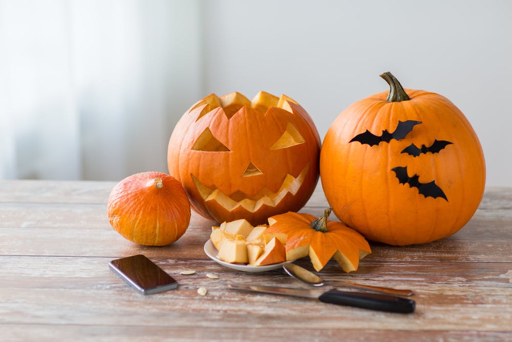 Un gros plan d'une table en bois couverte de décorations d'Halloween, de popcorn dans un bol motif toile d'araignée et de petits seaux en forme de citrouille, avec des personnes en costume floues à l'arrière-plan.