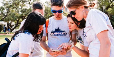 Five colleagues in matching grey event T-shirts huddle together outdoors on a sunny day, one holding a phone while a woman points at the screen and the others lean in to read it.