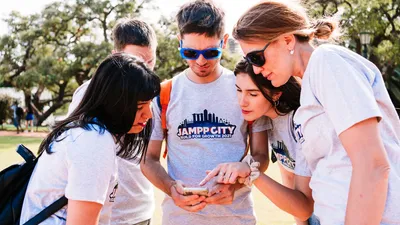 Five colleagues in matching grey event T-shirts huddle together outdoors on a sunny day, one holding a phone while a woman points at the screen and the others lean in to read it.