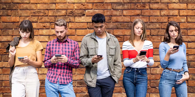 Un groupe diversifié de cinq jeunes adultes debout en rang devant un mur de briques texturé, chacun regardant attentivement l'écran de son smartphone.