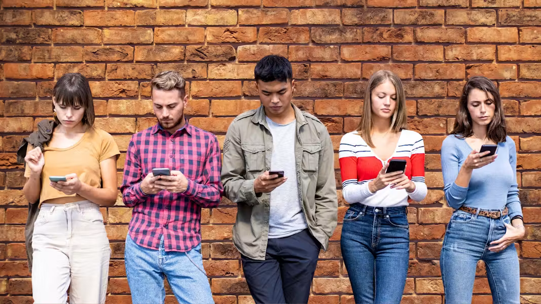 A diverse group of five young adults standing in a row against a textured brick wall, each looking down intently at their smartphone screen.