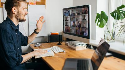 A side-view of a man wearing earphones, waving at his desktop monitor while hosting or participating in a large group video meeting from his home office desk.