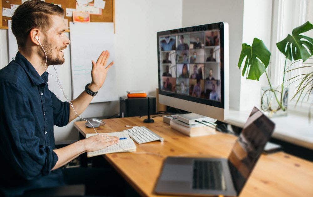 A side-view of a man wearing earphones, waving at his desktop monitor while hosting or participating in a large group video meeting from his home office desk.