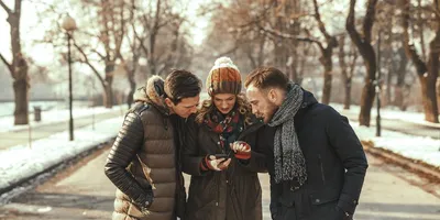 Drei Freunde in warmen Wintermänteln und Mützen stehen zusammen auf einem Parkweg und schauen gebannt auf ein Smartphone, das eine Frau in der Mitte in der Hand hält.